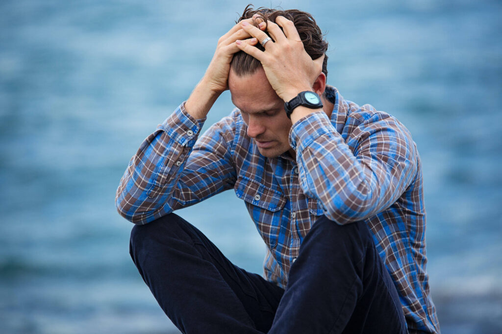 A person in a plaid shirt sits with hands on head, appearing distressed, against a blurred background of water. They wear a watch and seem contemplative.